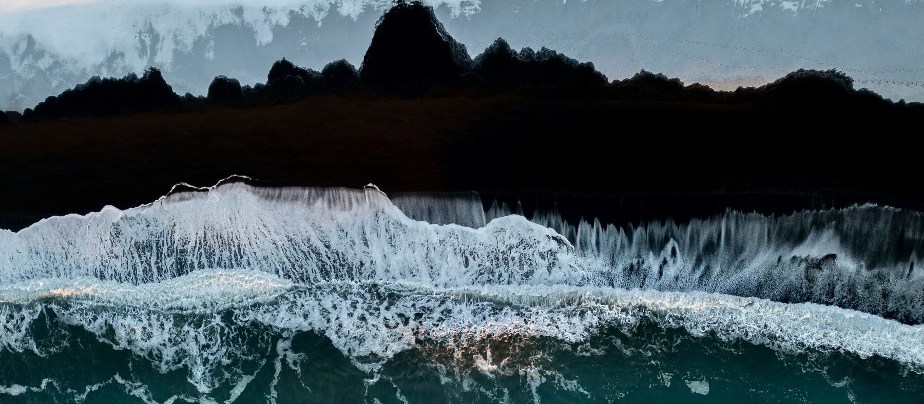 Waves breaking on a black sand shoreline from above